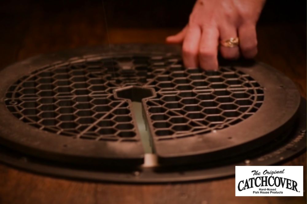 A person's hand is reaching into a Catch Cover Safety Grill Cover - CC09 on a wooden floor.