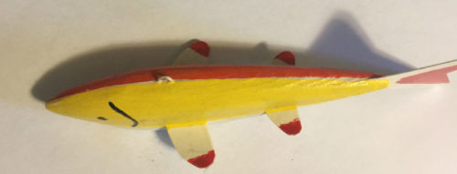 A yellow and red 6-1/2 Inch Lamont Mounsdon Wood Spearing Decoy fish is laying on a white surface.