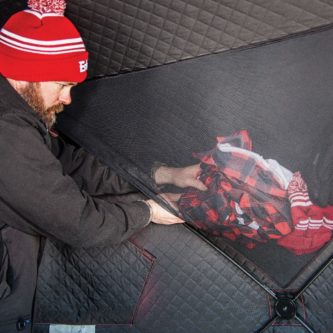 A man in a red beanie is putting clothes into an Eskimo Fatfish 949i HUB Ice Shelter.