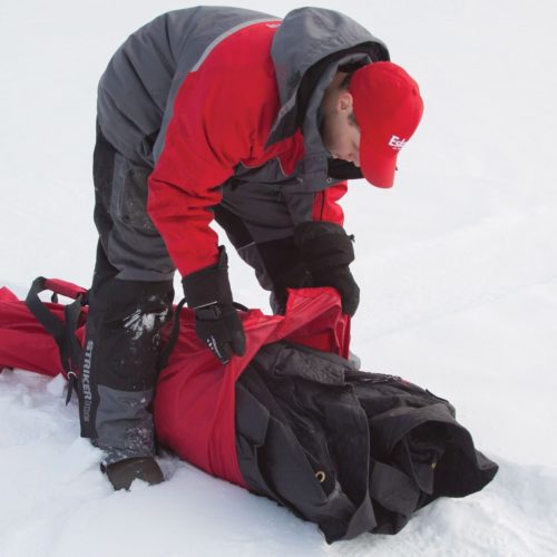A man in a red jacket is putting an Eskimo Fatfish 949i HUB Ice Shelter in the snow.