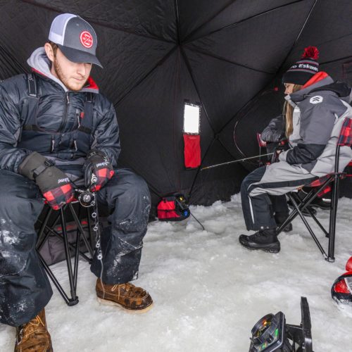 A man sits on a chair in an Eskimo 36150 QuickFish 6i Pop-Up Portable Insulated Ice Fishing Shelter, 6 Person.